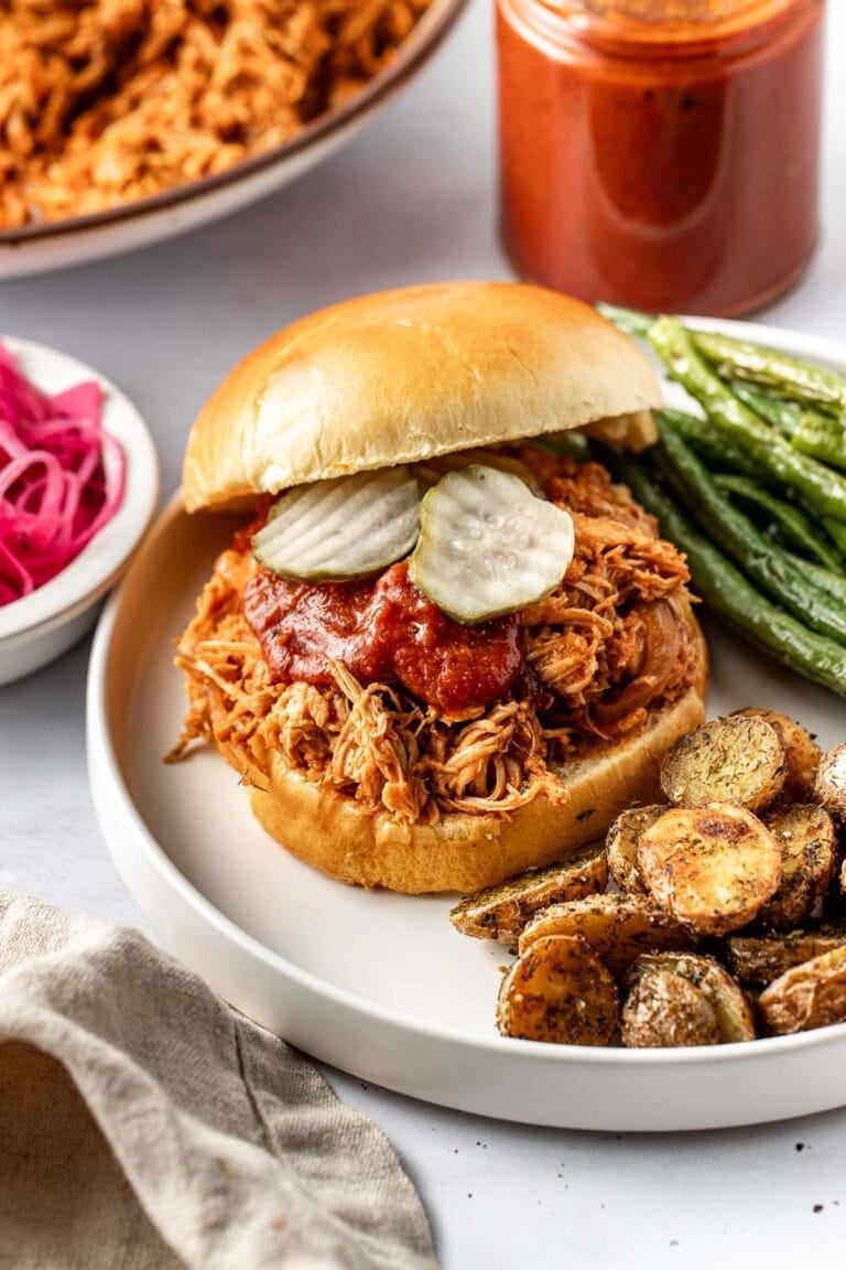 Close-up view of shredded BBQ chicken on a brioche bun topped with a drizzle of BBQ sauce and sliced pickles, with side dishes of roasted potatoes and green beans on the plate.