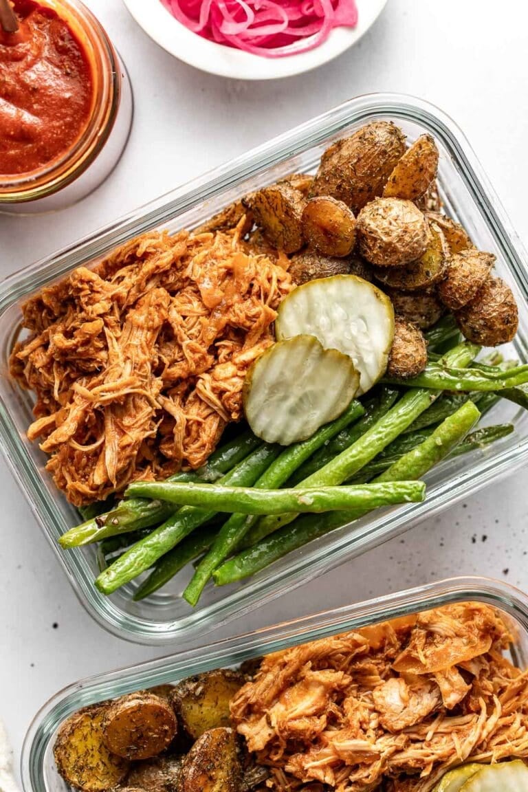 Overhead view of two glass meal prep containers filled with BBQ pulled chickenroasted potatoesand green beans topped with pickle slices.