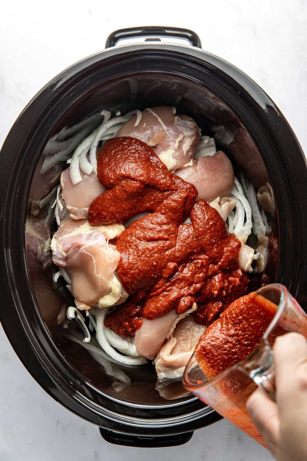 Overhead view of a slow cooker filled with chicken breasts, sliced white onions, and BBQ sauce being poured over the top.