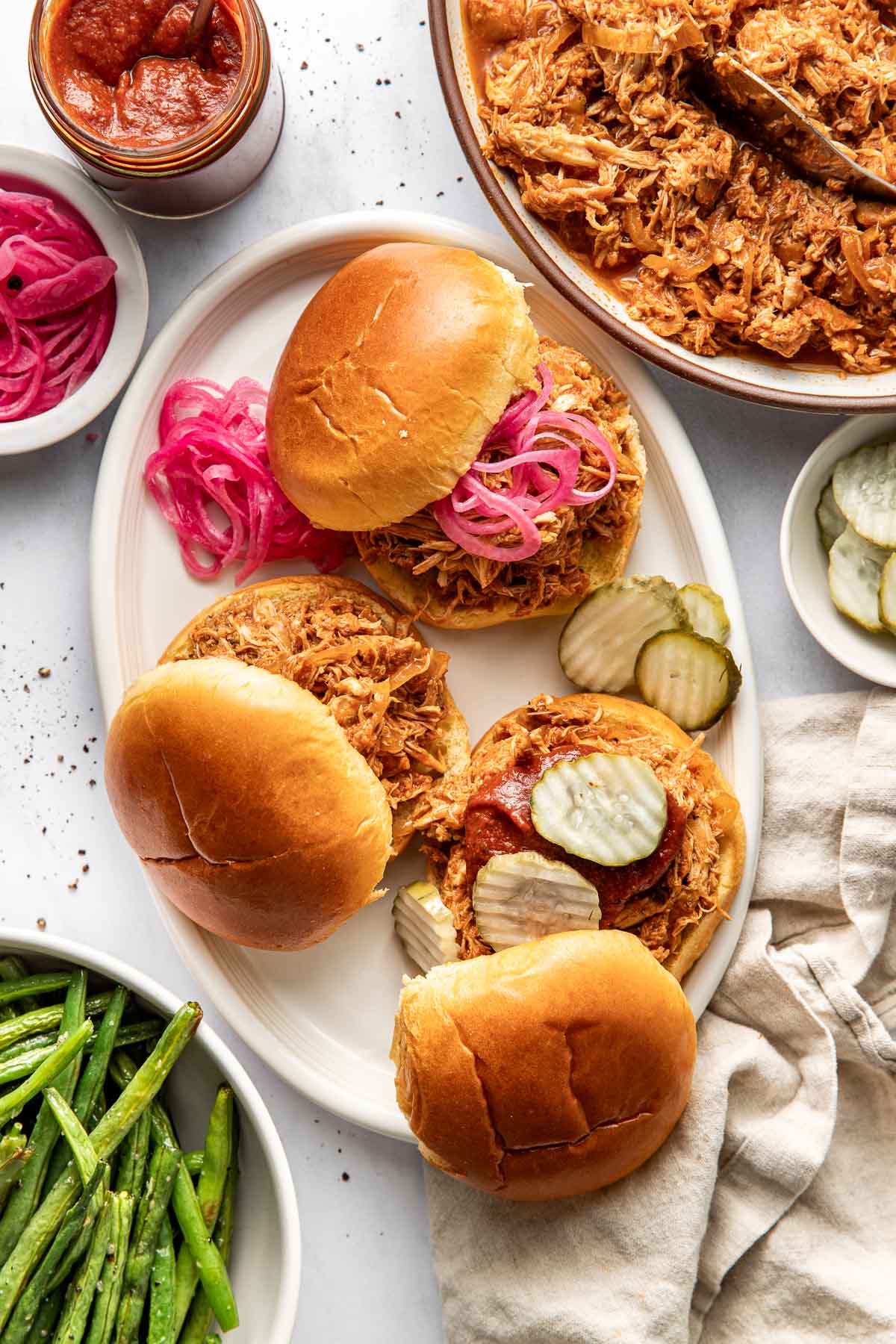 Overhead view of a tablescape showing BBQ Pulled Chicken sandwiches with different toppings including pickled onions, sliced pickles, and homemade BBQ sauce.