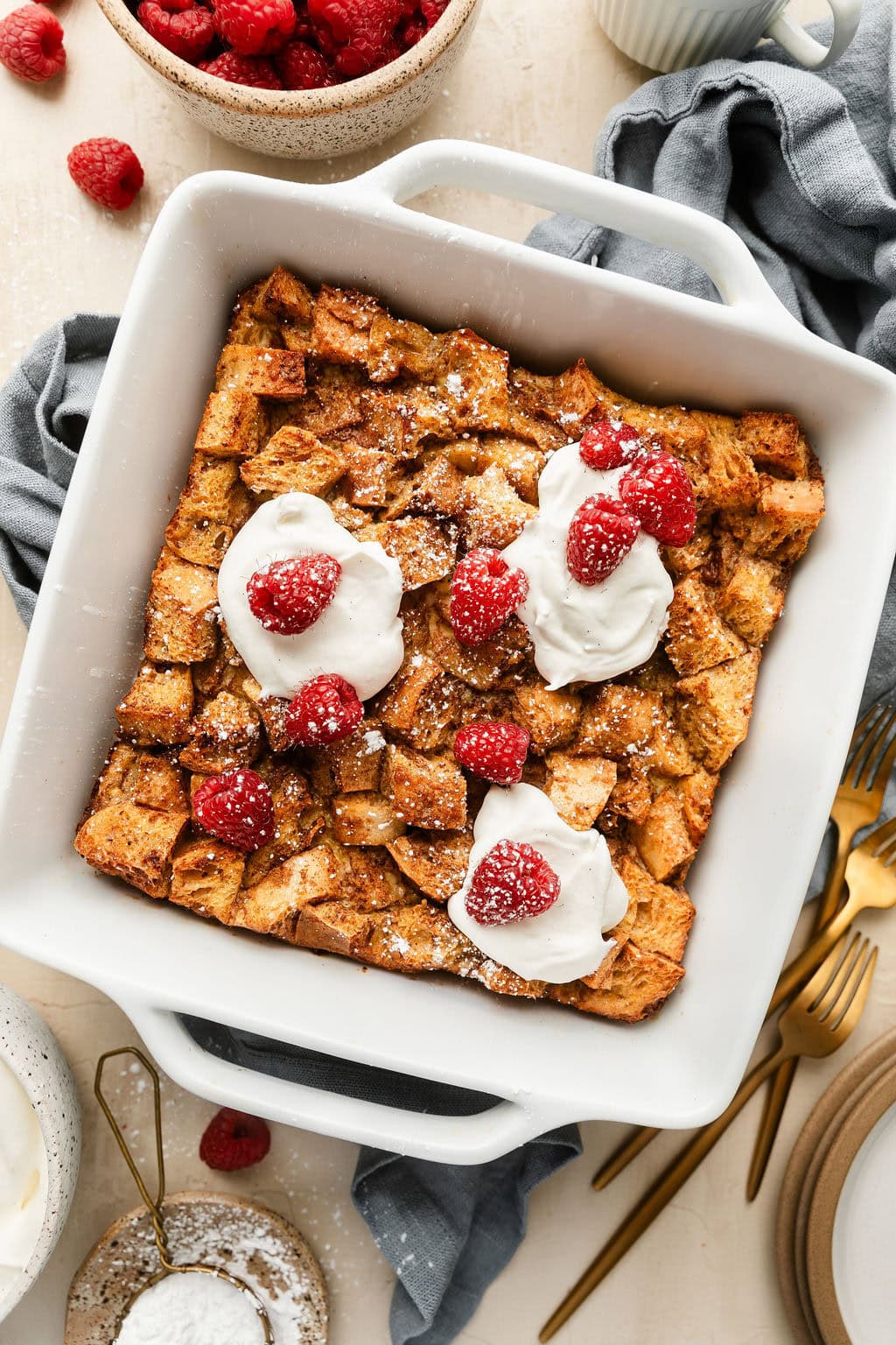Overhead view of a baking dish filled with freshly baked French Toast Casserole topped with raspberries, whipped cream, and sprinkled with powdered sugar. 