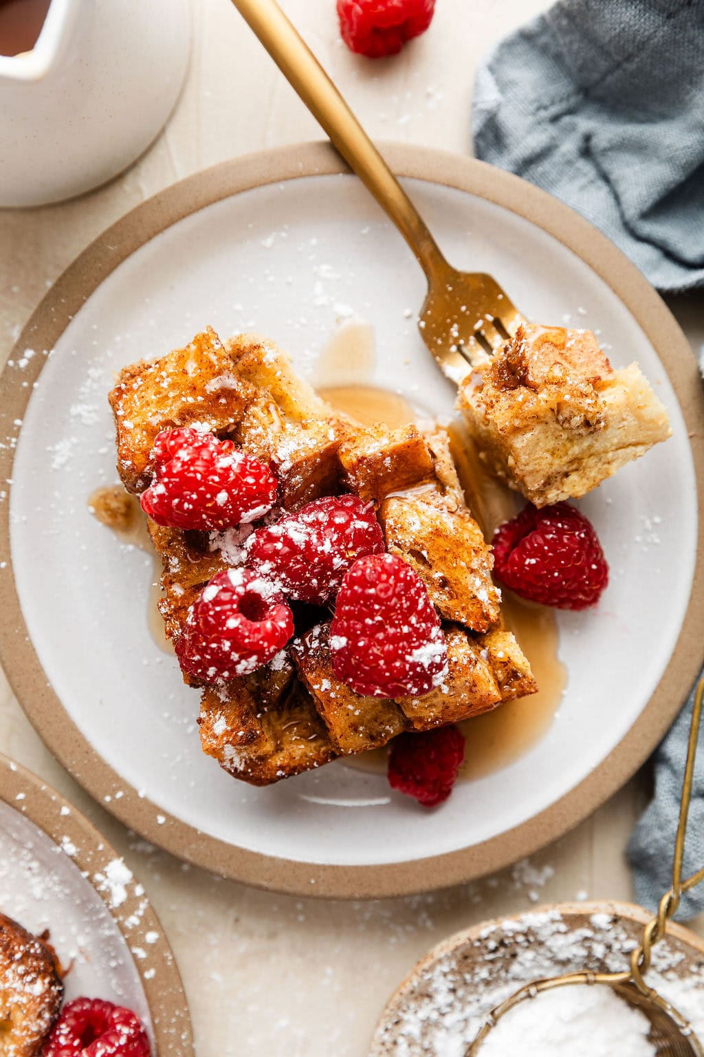 Overhead view of a plate filled with a piece of fresh toast casserole topped with raspberries, maple syrup and sprinkled with powdered sugar. 