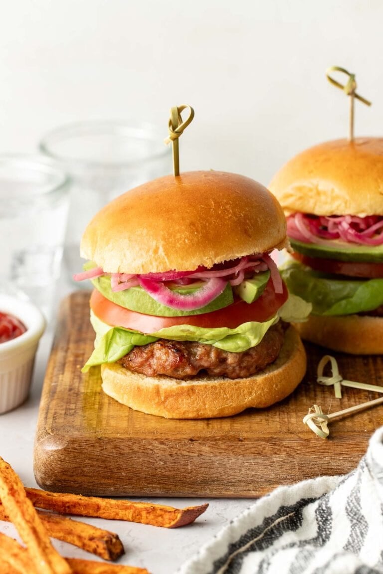 Close-up side view of two grilled turkey burgers on buns topped with slices of tomato, lettuce, avocado, and pickled red onions.