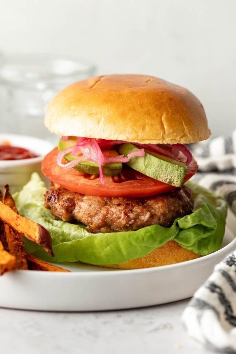 Close up view of a grilled turkey burger in a bun on a white plate over a leaf of romaine lettuce and topped with sliced tomato, avocado, and picked red onions.