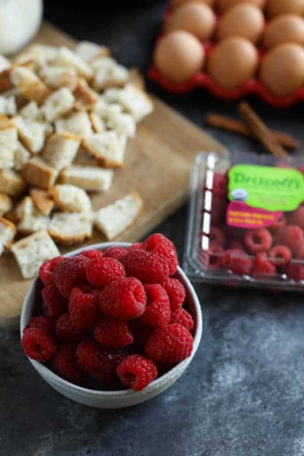 Cutting board with bread cubes, a dozen eggs and a bowl of raspberries on a table. 
