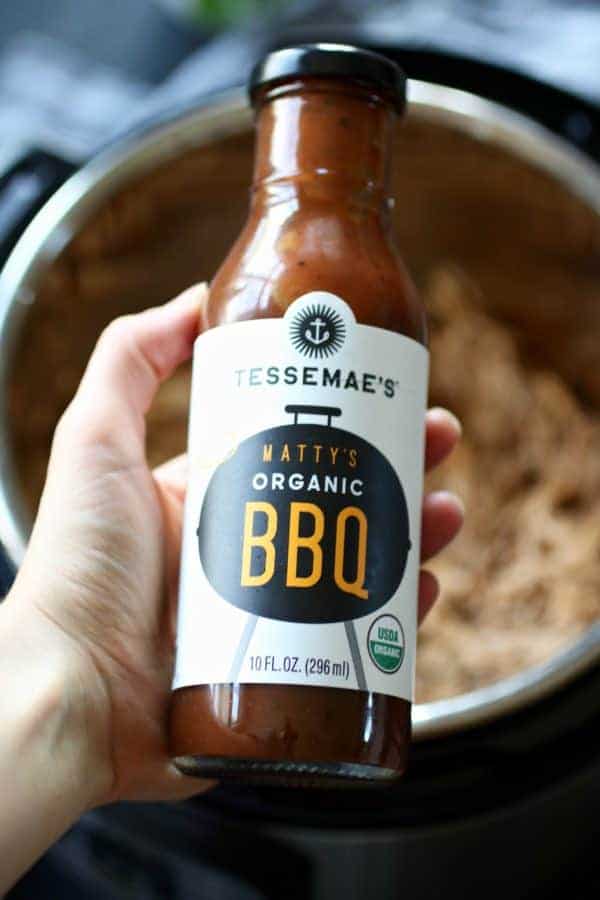 Tessemae's Organic BBQ Sauce held facing the camera with a bowl of Instant Pot Pulled Chicken blurred in the background