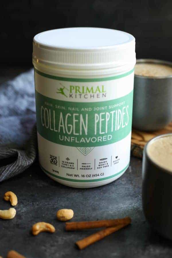 Canister of Primal Kitchen Collagen Peptides next to two mugs of Snickerdoodle Cashew Coffee with a few cashews and cinnamon sticks in the foreground. 