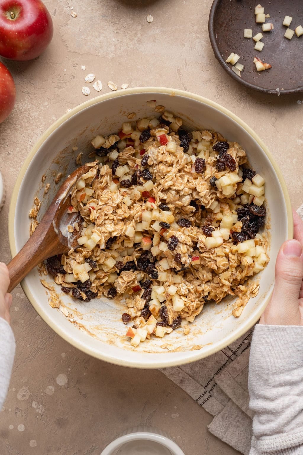 A hand using a wooden spoon to stir apple oatmeal cookie ingredients in a bowl.