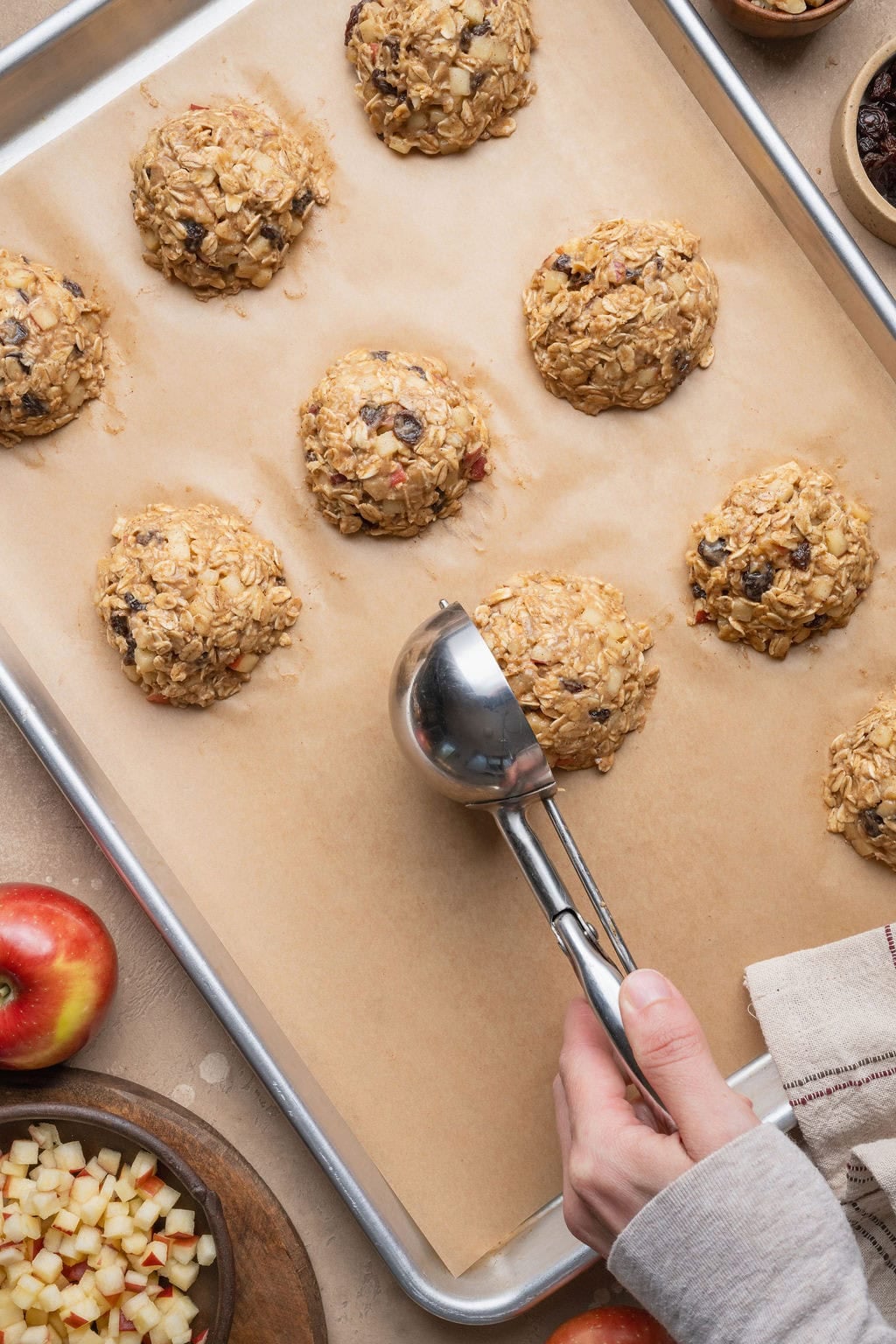 A hand using a cookie scoop to portion apple oatmeal cookies onto a parchment paper-lined baking sheet.