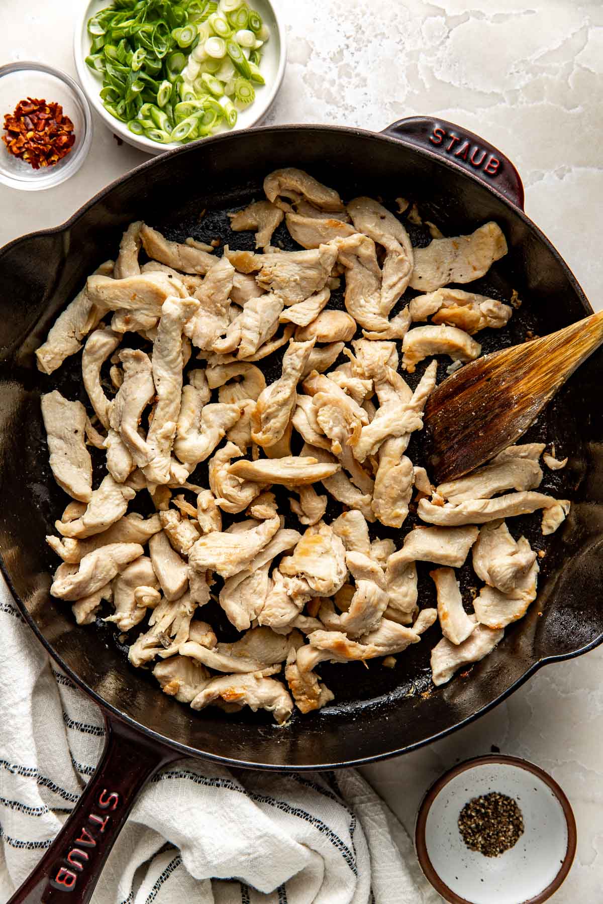 Overhead view of thinly sliced chicken in a skillet browning with a wooden spoon.