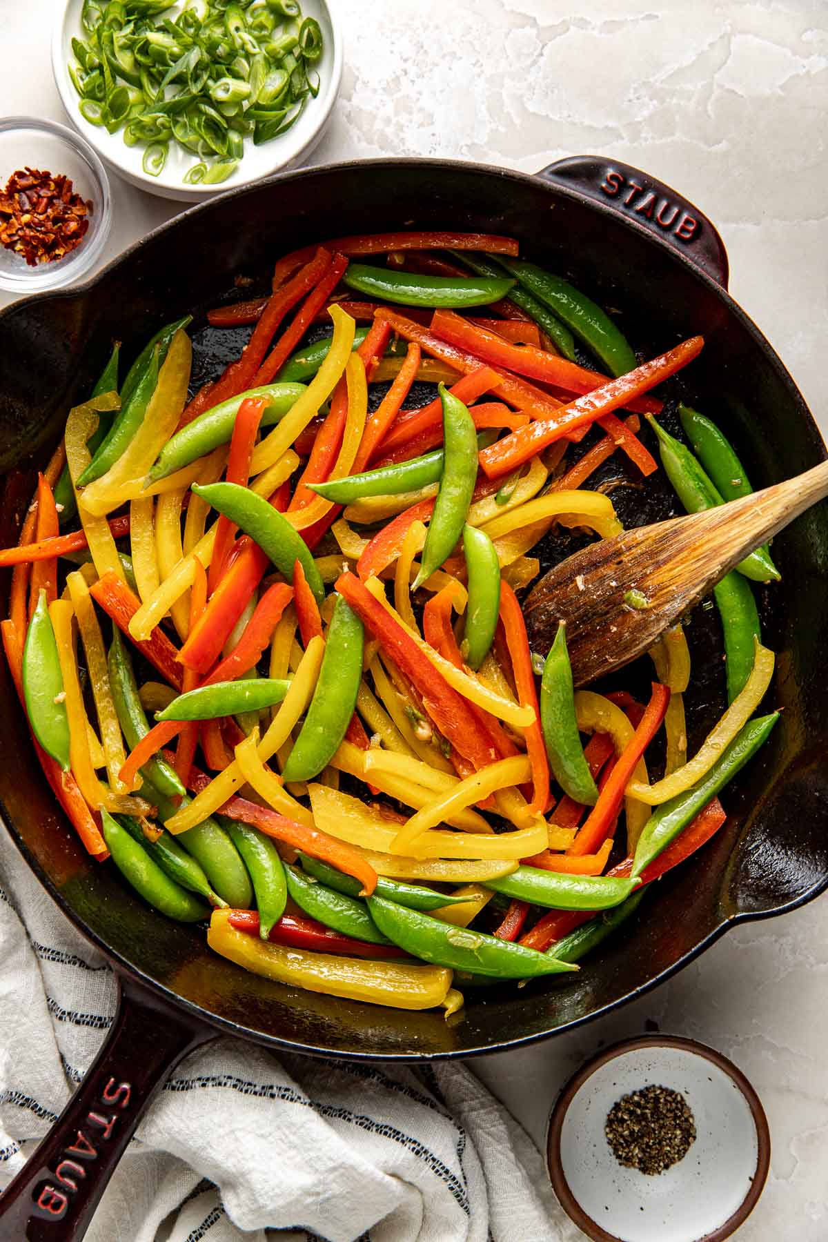 Overhead view of a black skillet filled with colorful sliced peppers and snap peas being stirred with a wooden spoon.