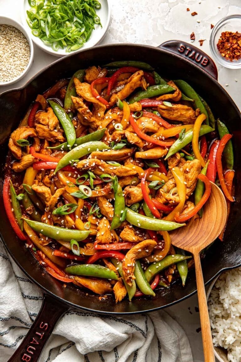 Overhead view of a cast iron pan of colorful chicken stir fry topped with green onions and white sesame seeds. 