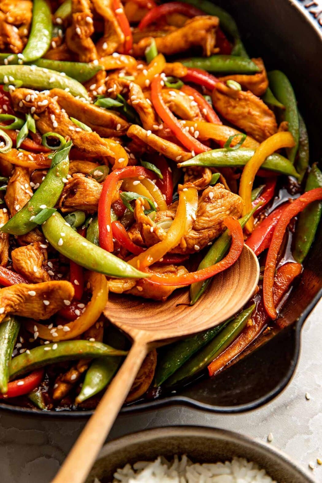 Close up view of a skillet of Chicken Stir Fry being stirred with a wooden spoon topped with sesame seeds.