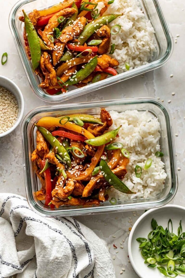 Overhead view of two glass meal prep containers filled with chicken stir fry and white rice sprinkled with sesame seeds and green onions.