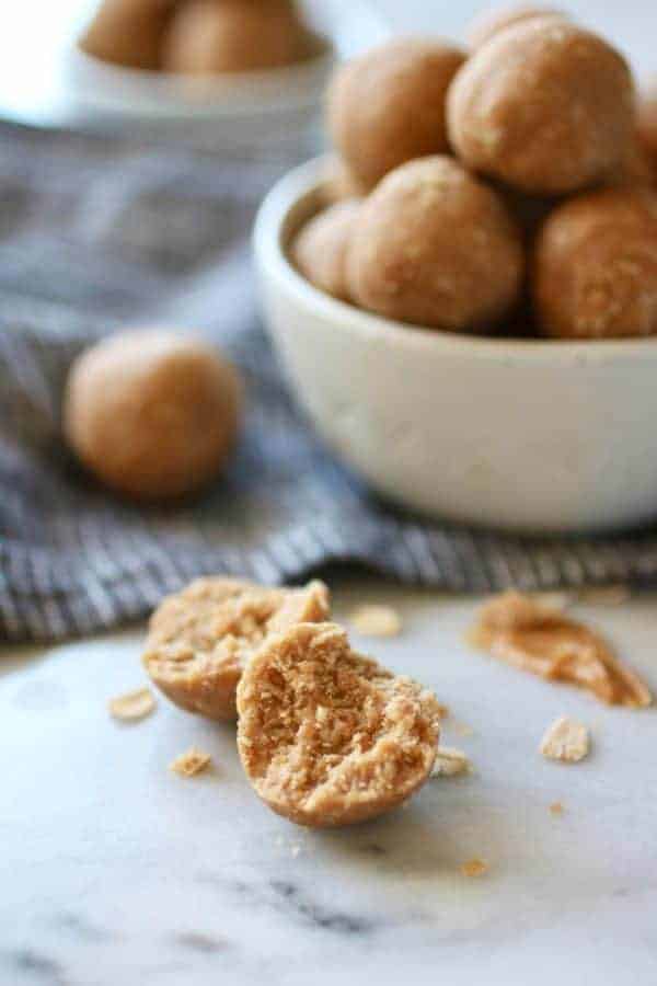 A 3-Ingredient Peanut Butter Bite split in half on a table in front of a bowl of 3-Ingredients Peanut Butter Bites