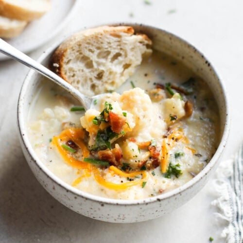 A warm bowl of Loaded Cauliflower Soup topped with shredded cheese, herbs and bacon, serves with yummy bread.