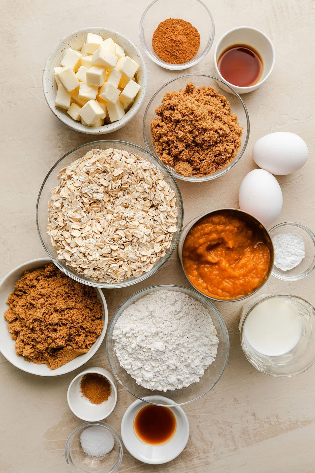 Overhead view of a variety of ingredients for pumpkin pie bars in different sized bowls with two eggs included on the side.