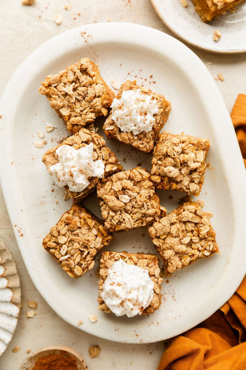 Overhead view of a white platter filled with pumpkin pie bars with some of the squares topped with a dollop of whipped cream and sprinkled with pumpkin pie spices.