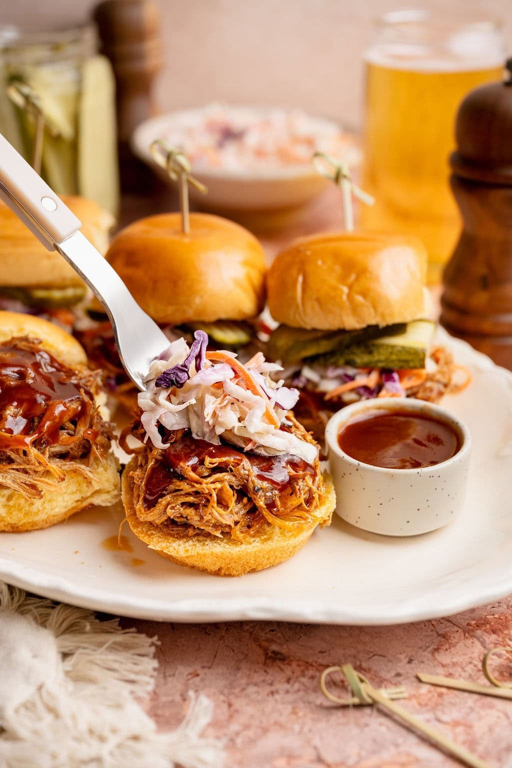 Close up view of a platter of Pulled Pork Sliders with a fork adding coleslaw to the top of one of the sandwiches