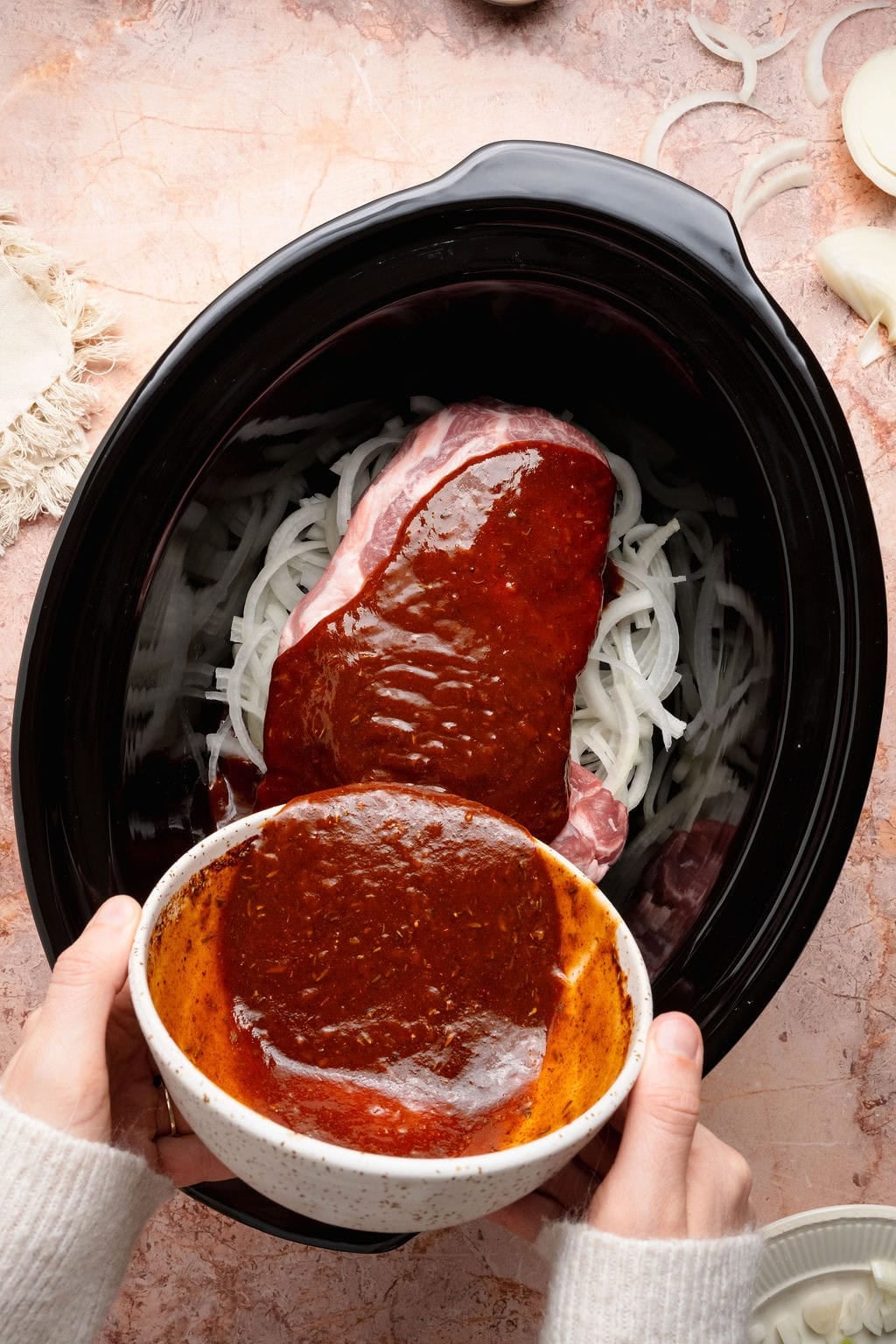 Overhead view of a slow cooker with a pork roast and onions inside with hands pouring a BBQ sauce mixture on the top.