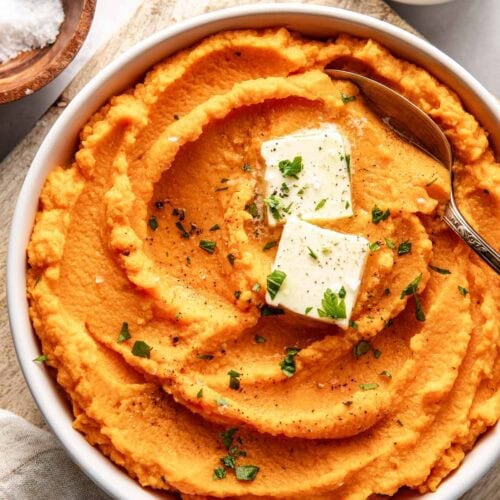 Overhead view of a bowl of vibrant orange Mashed Sweet Potatoes topped with butter and fresh herbs.
