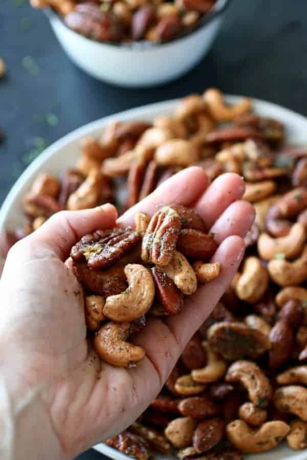 Ranch Roasted Mixed Nuts being held in a hand. Bowl and plate of nuts in the background. Photo taken on a dark background.