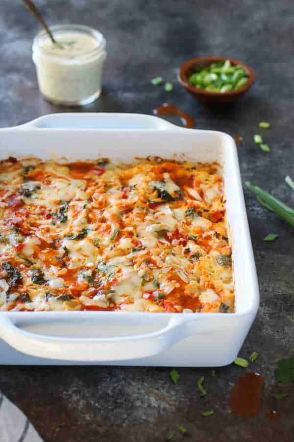 Front angle photo of Buffalo Chicken Bake in a white 9x9 casserole dish with a small mason jar of ranch and small wooden bowl with fresh chives in the background.