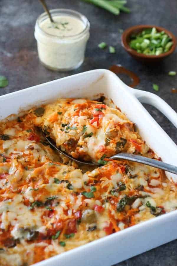 side angel photo of Buffalo Chicken Bake in a 9x9 white dish with a silver spoon holding a scoop. Small mason jar filled with ranch and a small wooden bowl of chives in the background. 