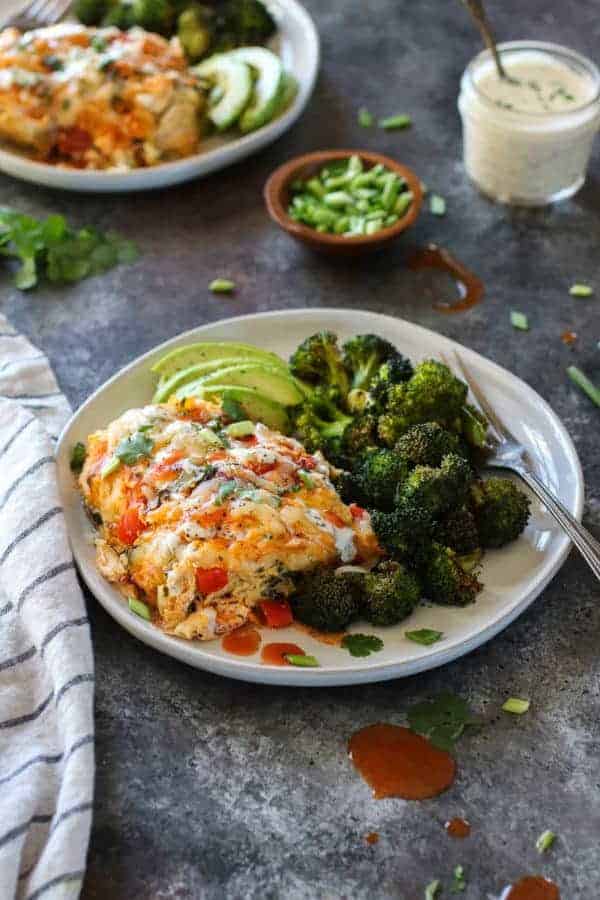 Side angle photo of Buffalo Chicken Bake on white plate with roasted broccoli and sliced avocado. Drizzled with ranch and buffalo sauce.