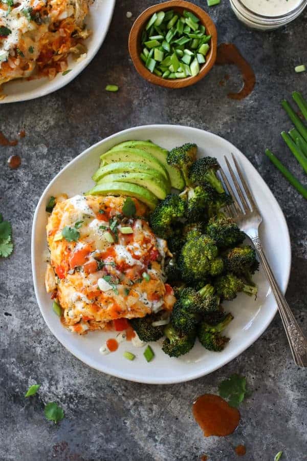 Top view photo of Buffalo Chicken Bake on a white plate with a side of roasted broccoli and sliced avocado. Drizzled with ranch and buffalo sauce. 