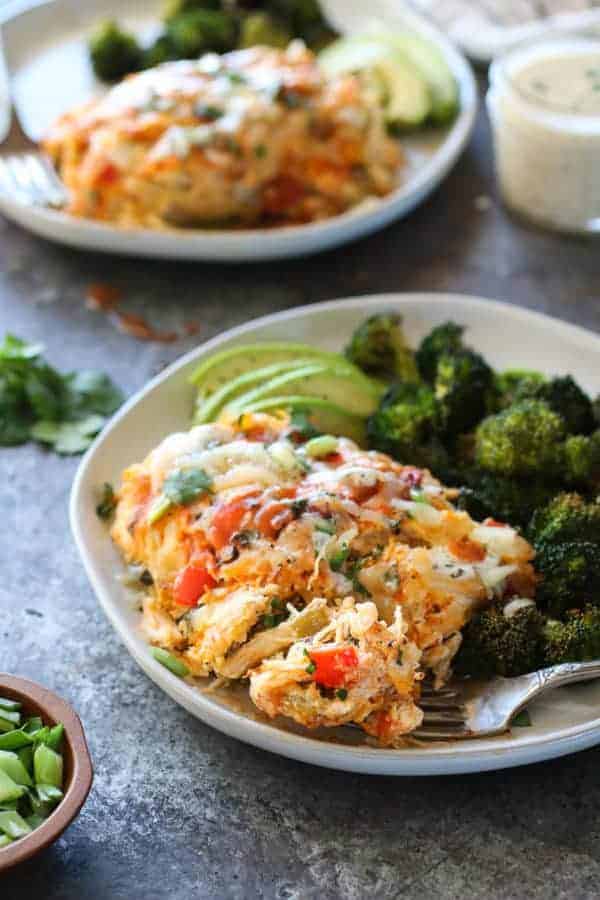 Side angle photo of Buffalo Chicken Bake on white plate with roasted broccoli and sliced avocado. Drizzled with ranch and buffalo sauce.