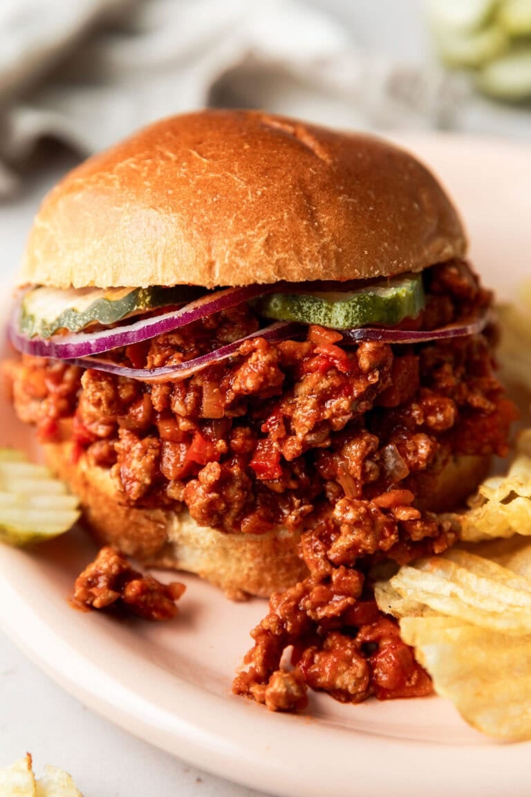 Close up view of a plate of chips and a turkey sloppy joe on a bun topped with pickles and red sliced onions. 