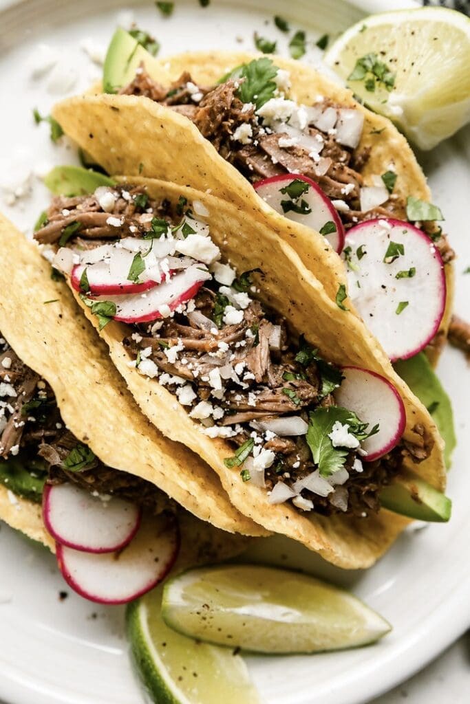Close up view slow cooker beef barbacoa in hard shell tacos with sliced radishes, fresh cilantro, and sprinkle of white cheese.