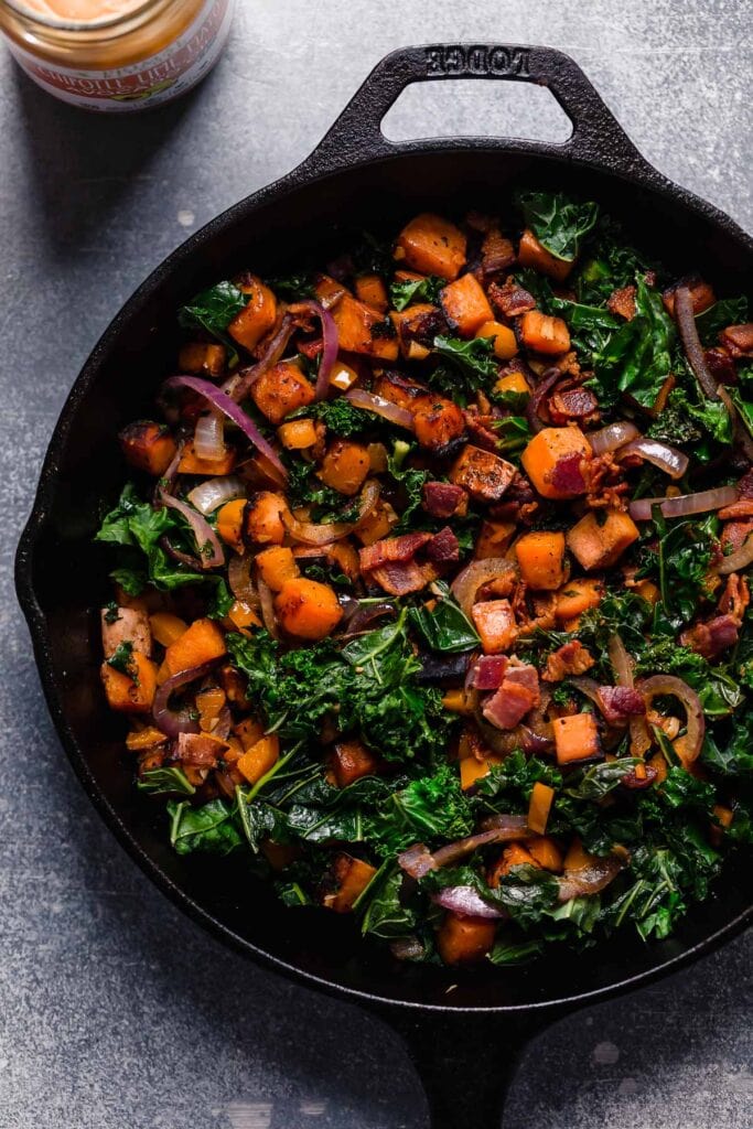 An overhead view of sautéed sweet potato, kale, red onion, and bacon in a black cast iron skillet. 