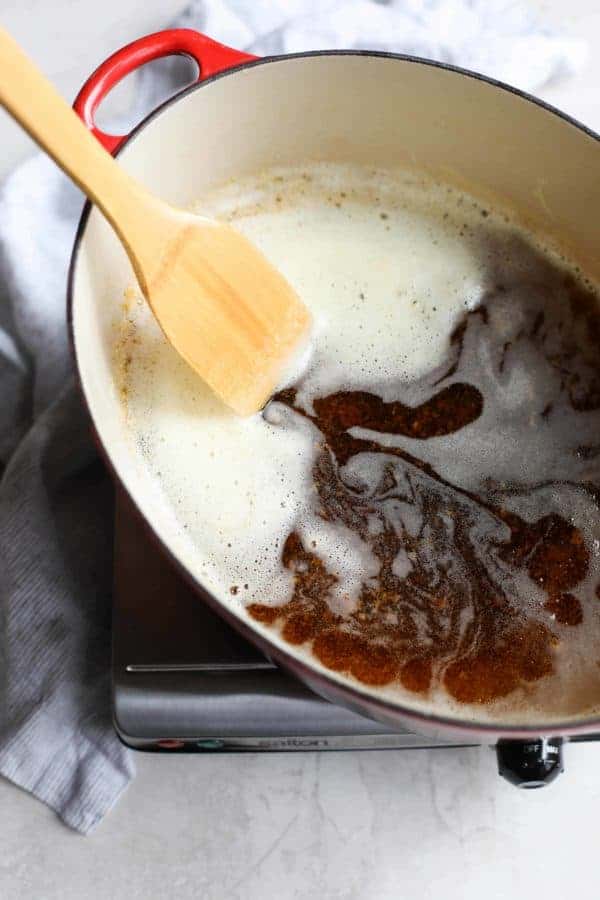 Finished product - ghee in a Dutch oven with spatula pulling aside the foam to reveal the clarified liquid and brown solids at the bottom of the pan. 