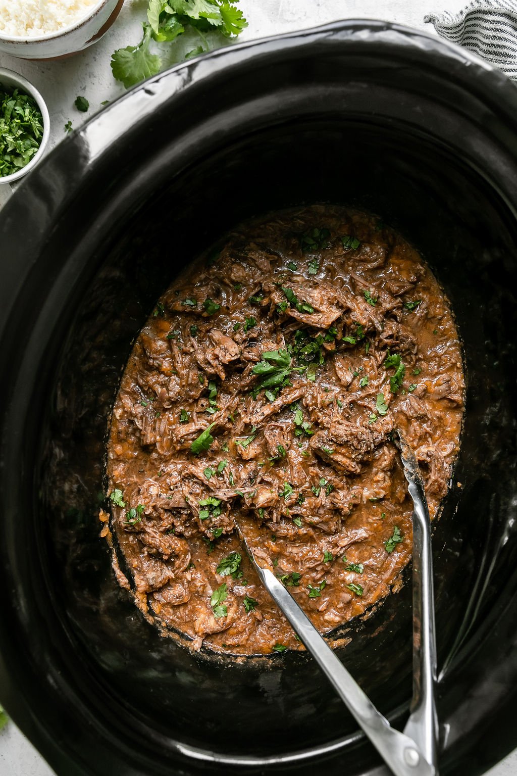 Shredded saucy beef barbacoa in black crockpot with tongs.