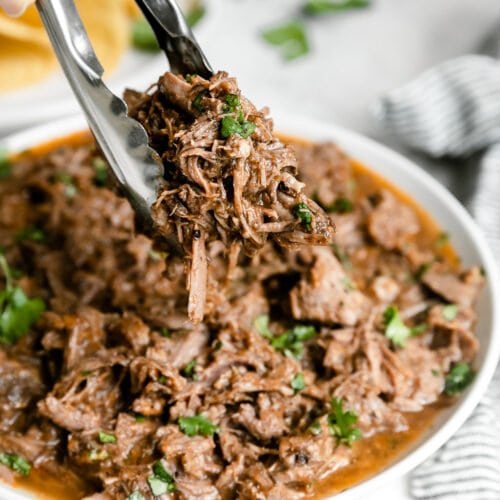 Shredded saucy slow cooker beef barbacoa in white bowl with tongs lifting serving up.