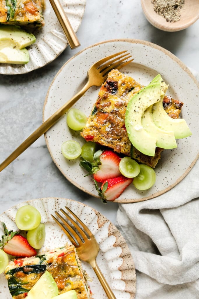 Serving of sweet potato breakfast casserole on plate topped with avocado slices, fresh fruit on side.