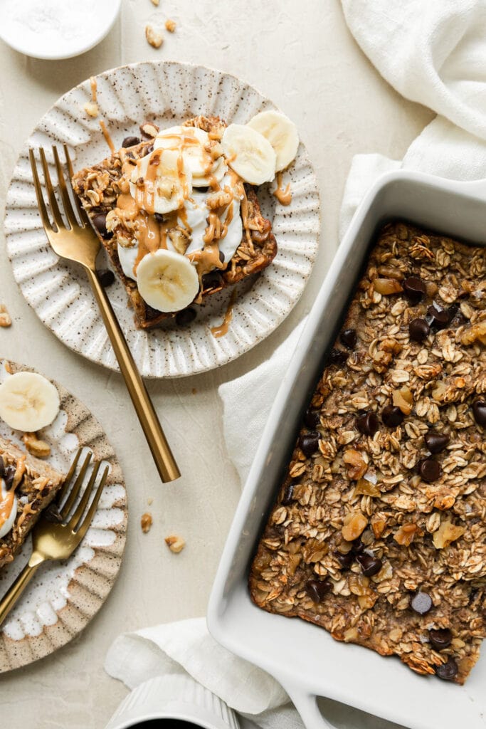 Banana baked oatmeal serving on a plate topped with yogurt, nut butter, and banana slices, baking dish with baked oatmeal to the side.