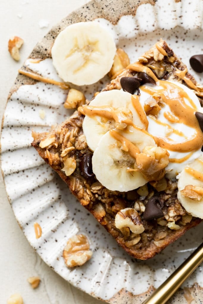 Close up overhead view banana baked oatmeal with chocolate chips on stone plate topped with yogurt, nut butter and banana slices. 