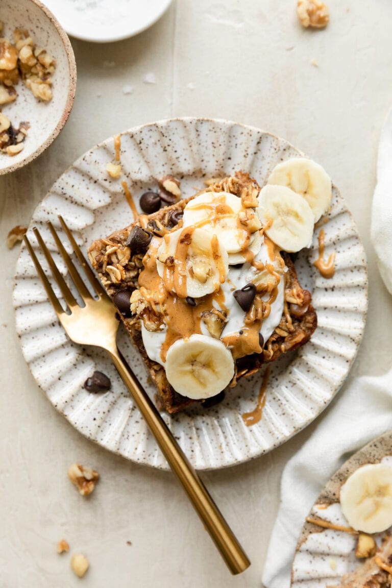 Overhead view banana chocolate chip baked oatmeal serving on scalloped edge plate topped with yogurt, banana slices, and peanut butter drizzled over top.