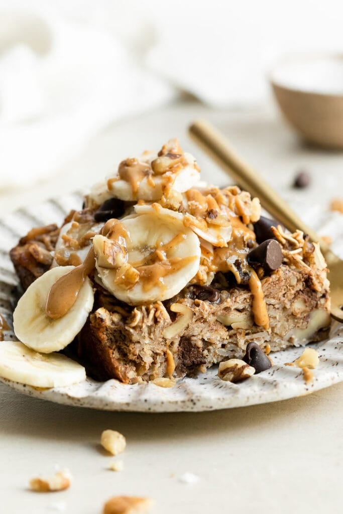 Close up side view banana baked oatmeal with chocolate chips and walnuts plated on stone plate topped with nut butter and banana slices. 