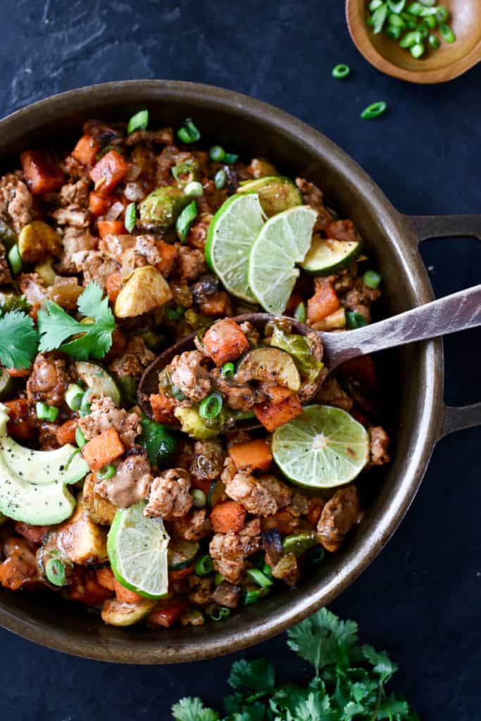 Iron skillet with cooked ground turkey, sweet potato, zucchini, brussels sprouts, onion with lime, avocado and onion garnish