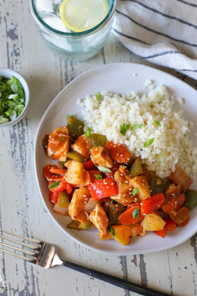 Sweet and Sour chicken with peppers and a side of cauliflower rice on white distressed background with a bowl of green onions and glass of lemon water. 4 of 10 Quick and Easy Dinner Ideas.