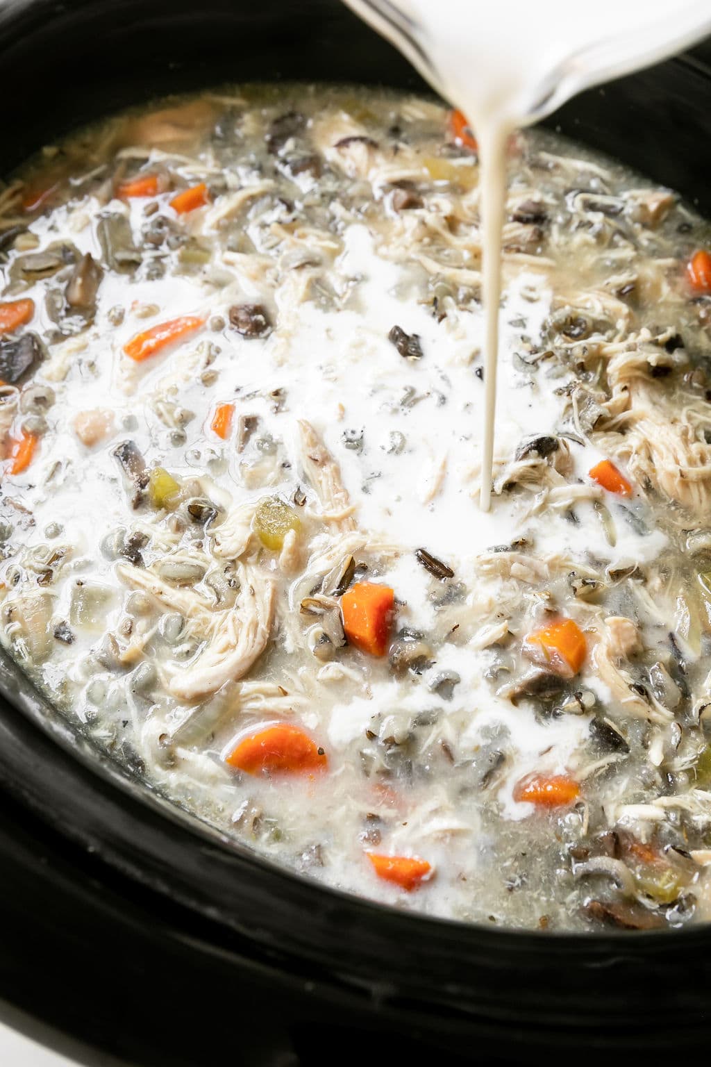 Cream being poured into a black crockpot filled with creamy chicken wild rice soup.