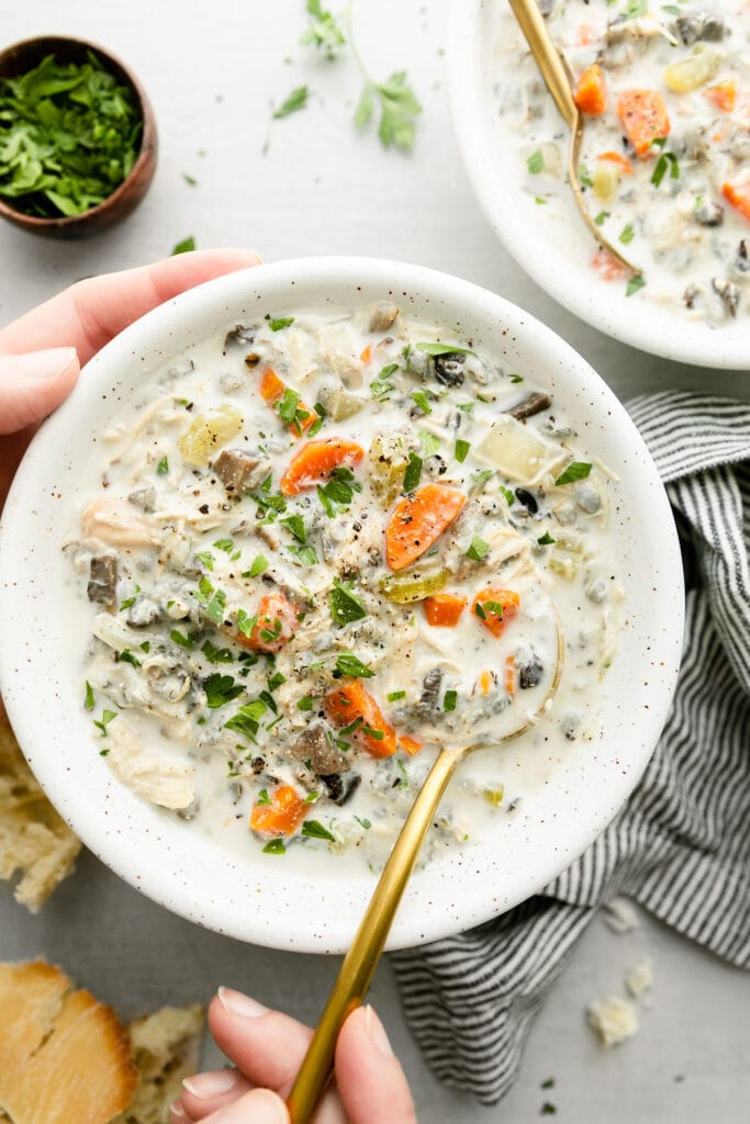 Two hands holding a bowl filled with creamy chicken wild rice soup, sprinkled with black pepper and fresh herbs.