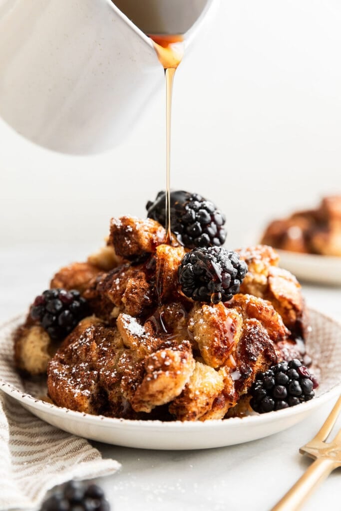 Maple syrup being poured over top of french toast casserole serving on plate, topped with blackberries and powdered sugar