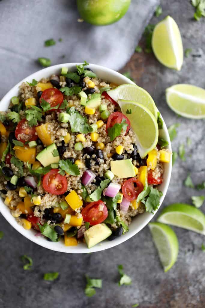Colorful quinoa salad with avocado, peppers, tomatoes, onions, limes and black beans in bowl surround by lime wedges