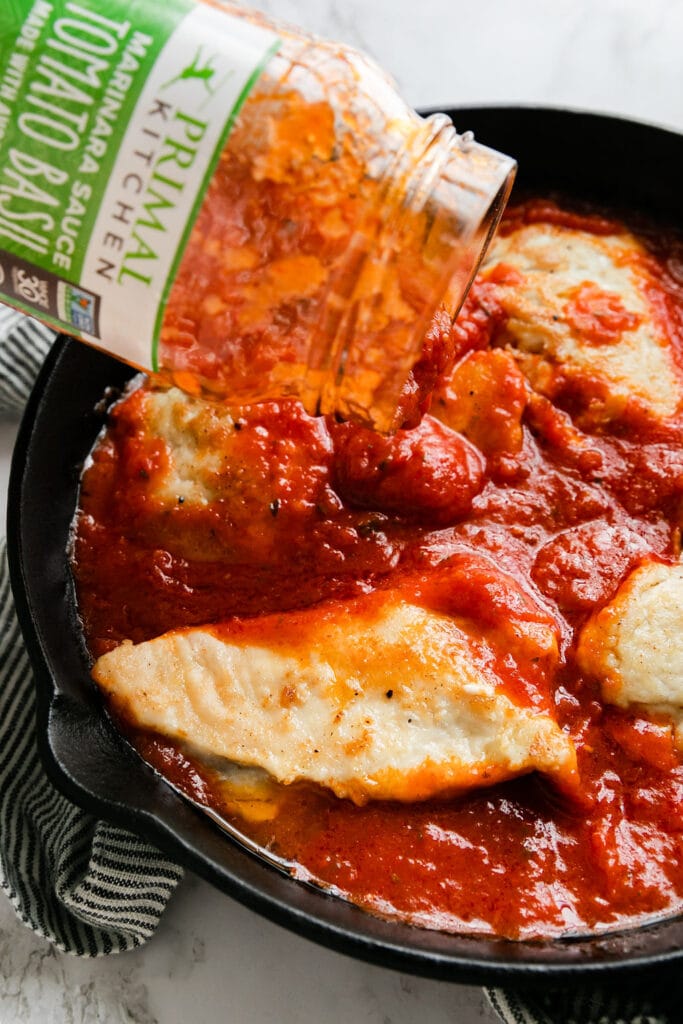 A jar of tomato basil marinara sauce being poured over cooked chicken breast in cast iron skillet.