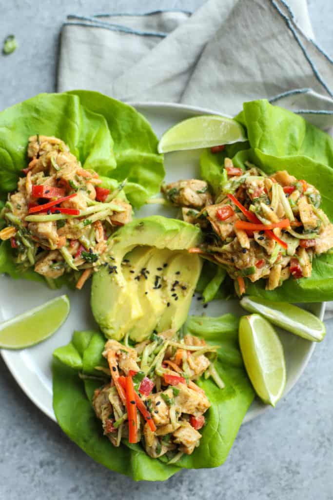 Three lettuce cups on a plate filled with chicken salad containing peppers, broccoli slaw and avocado slices in the middle and lime garnishes